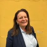 En kvinne med brunt går smiler til kamera foran en gul vegg.