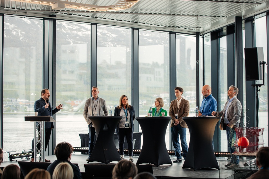 Paneldebatt med seks deltakere på scenen og en moderator, foran store vinduer med utsikt mot fjord og fjell