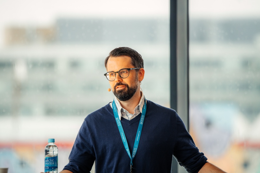 Person med mikrofon og navneskilt snakker under et arrangement, med en vannflaske på bordet foran.