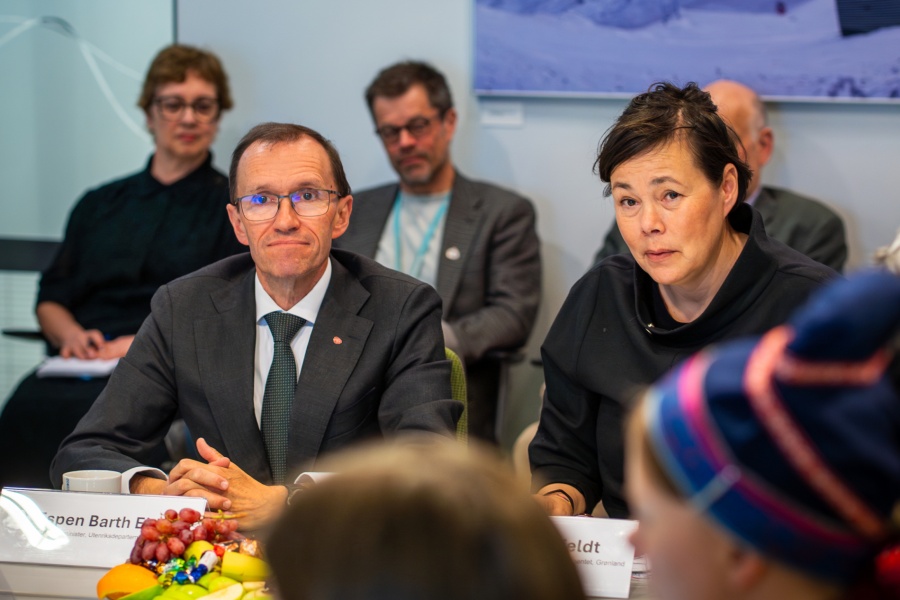 To møtedeltakere sitter ved et bord med navneskilt og frukt, mens flere andre følger med i bakgrunnen under et formelt møte.