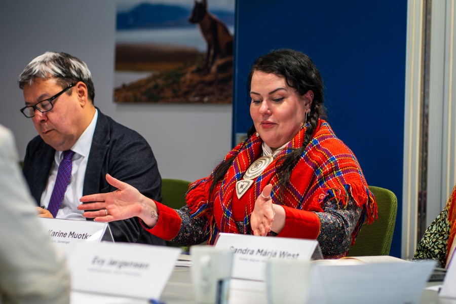 En kvinne snakker engasjert under et møte, mens en mann sitter ved siden av.