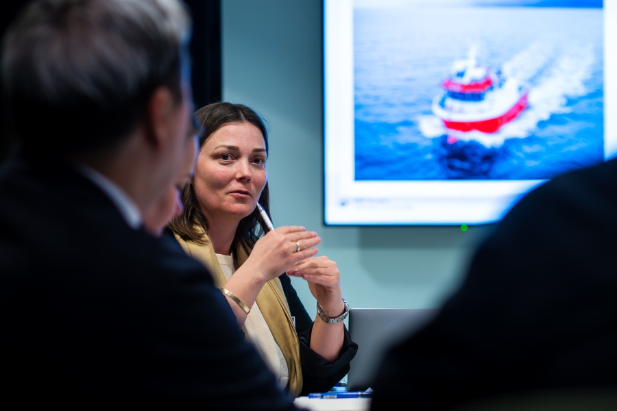 En kvinne snakker i et møte mens hun holder en penn. I bakgrunnen vises et bilde av en båt på en skjerm.