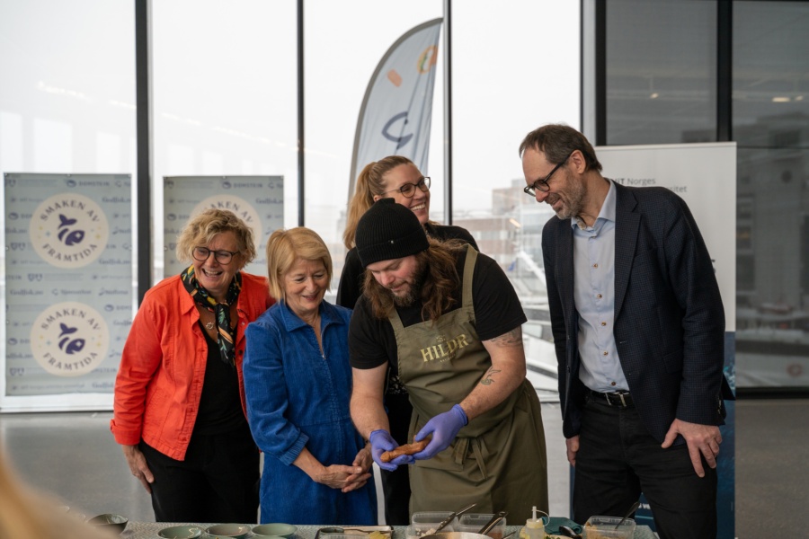 En kokk viser frem mat til en gruppe smilende personer under et arrangement, med bannere i bakgrunnen.