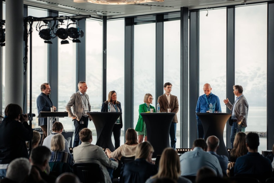 Syv paneldeltakere står samlet bak høye bord på en scene under en konferanse, med et publikum foran seg. Store vinduer i bakgrunnen viser snødekte fjell og hav. En deltaker til høyre snakker mens de andre lytter.