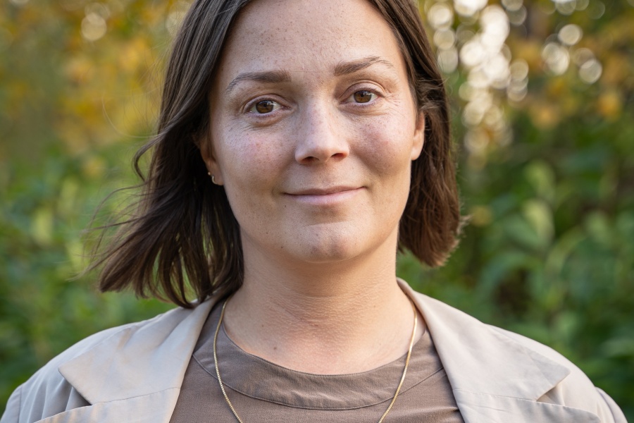 Portrett av en kvinne utendørs, med kort brunt hår, beige jakke og brun topp, foran grønn og gyllen høstlig bakgrunn.