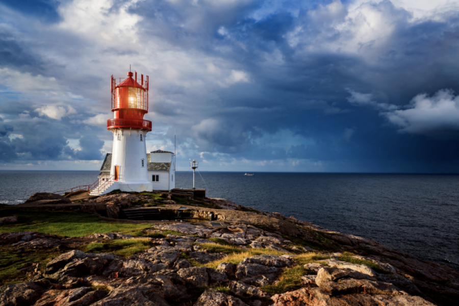 Fyrtårn med hvit base og rød topp står på en klippe ved havet, med dramatisk himmel og skyer i bakgrunnen.