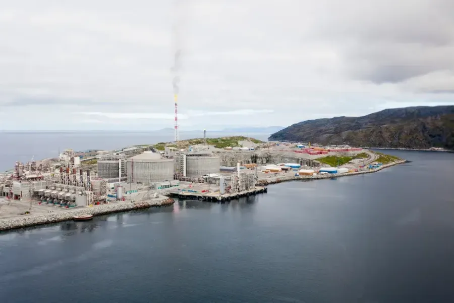 Oversiktsbilde av et stort industrikompleks ved kysten, med røyksøyle fra en pipe og fjell i bakgrunnen.