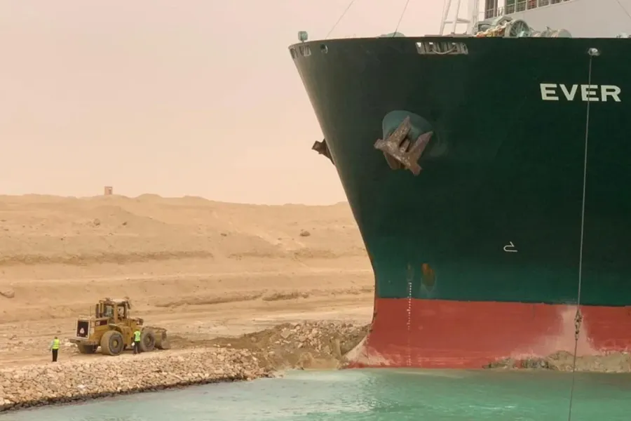 Et stort containerskip sitter fast på grunn i en kanal, mens en gravemaskin og arbeidere står ved siden av for å prøve å frigjøre det.