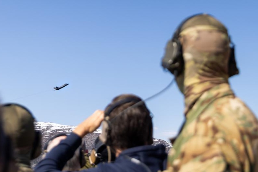 oldater med hørselvern og kamuflasjeuniform ser på et jagerfly som flyr over et fjellområde under en militær øvelse.