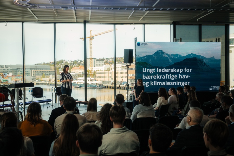 En kvinne står og holder innlegg foran et publikum i et lyst lokale med store vinduer mot havna i Tromsø. På en stor skjerm ved siden av henne står teksten «Ungt lederskap for bærekraftige hav- og klimaløsninger», med logoen til Senter for hav og Arktis. Publikum følger med på presentasjonen, og bakgrunnen viser bygg, båter og en kran ved havna.