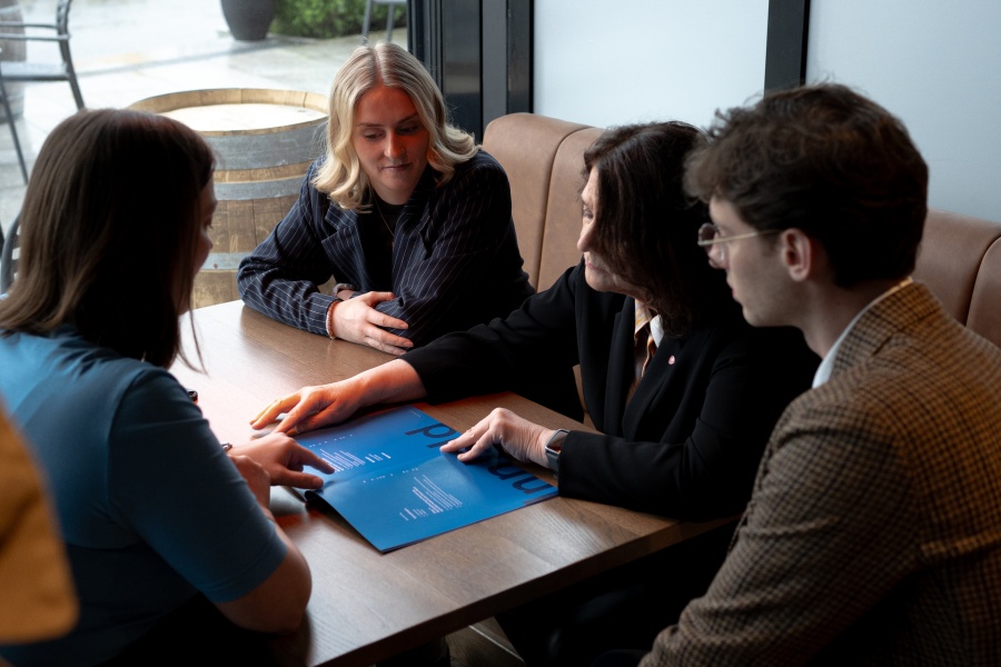  Fire personer sitter rundt et bord i en samtale. De ser på et blått dokument som ligger foran dem.