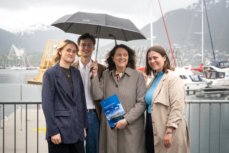 Fire personer står sammen utendørs ved en marina på en regnværsdag. En av dem holder en paraply, og en annen holder en rapport om marine næringsparker. Fjell og båter er synlige i bakgrunnen.