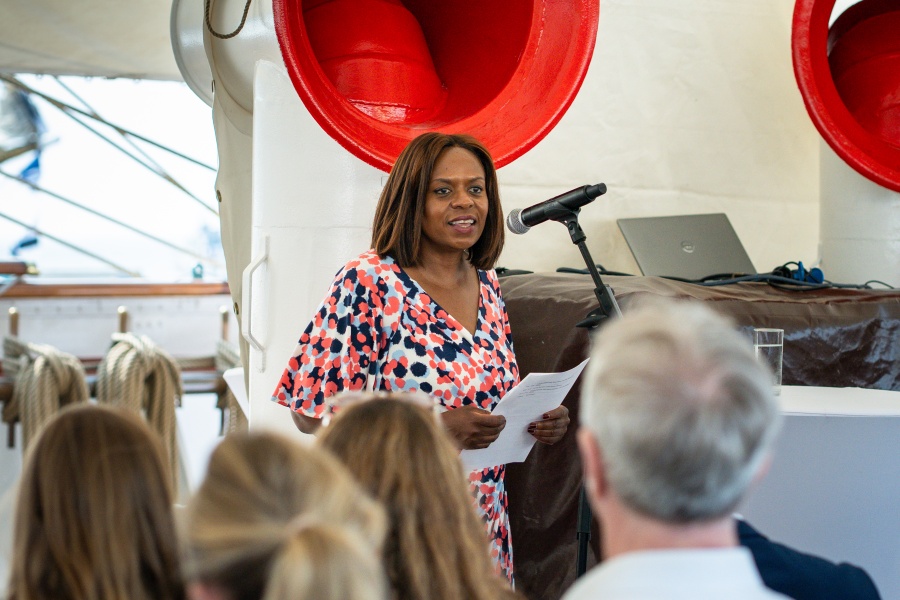 En kvinne står foran en mikrofon og holder et innlegg for et publikum. Hun holder ark i hånden og står i et maritimt miljø.