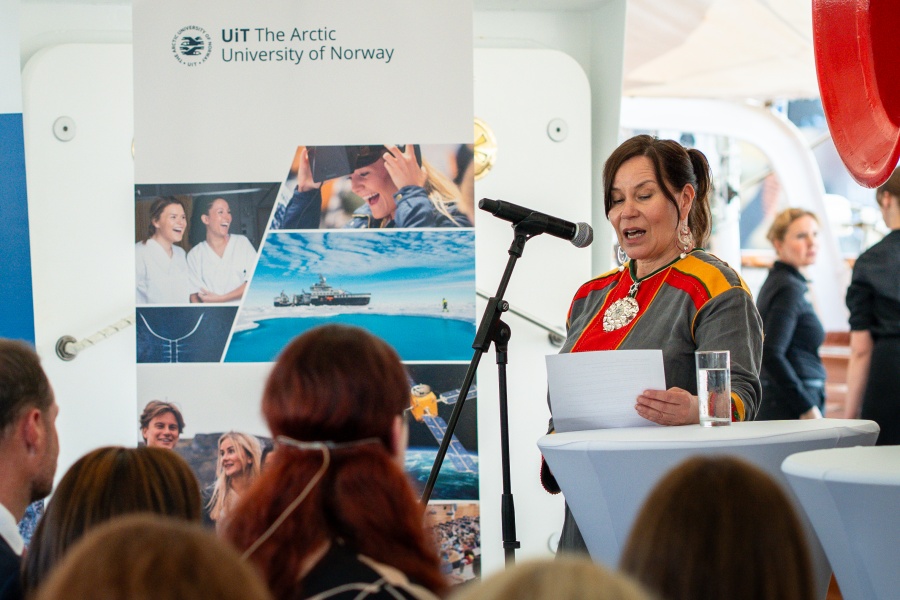 En kvinne i en rød, grå og oransje kofte står foran en mikrofon og taler til en forsamling. 