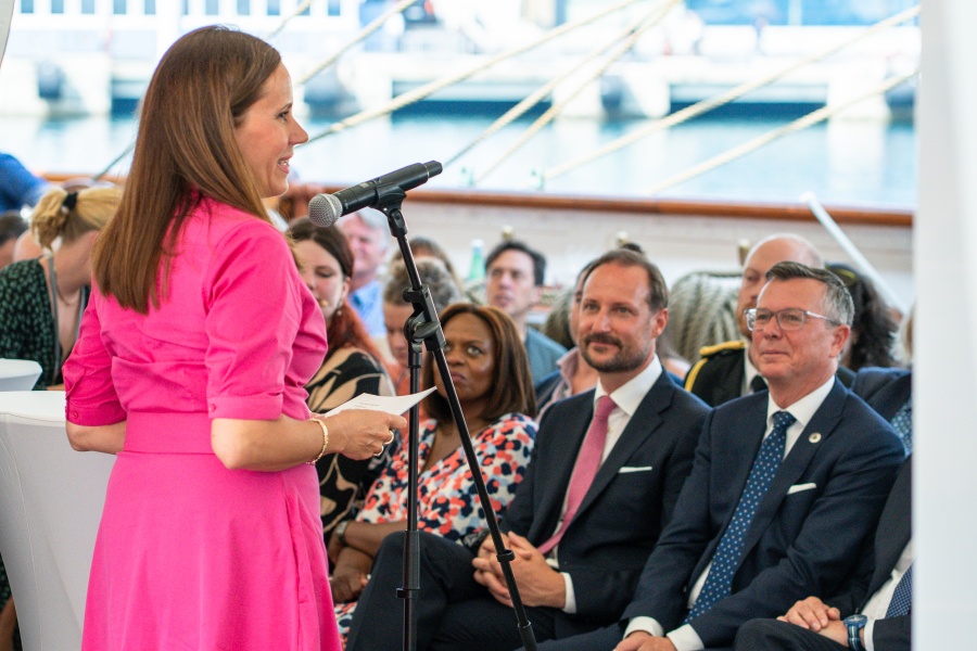 En kvinne i rosa kjole snakker i mikrofon foran et publikum på et arrangement. Flere personer sitter og lytter i bakgrunnen.