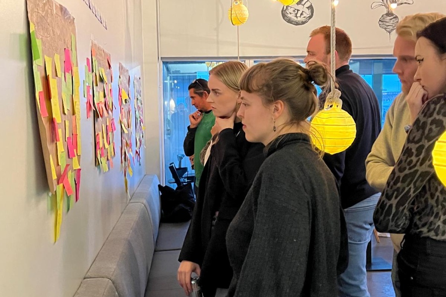 En gruppe deltakere står samlet foran en vegg med store ark dekket av fargerike post-it-lapper, og diskuterer ideer i en workshop-setting. Stemningen er konsentrert og engasjert