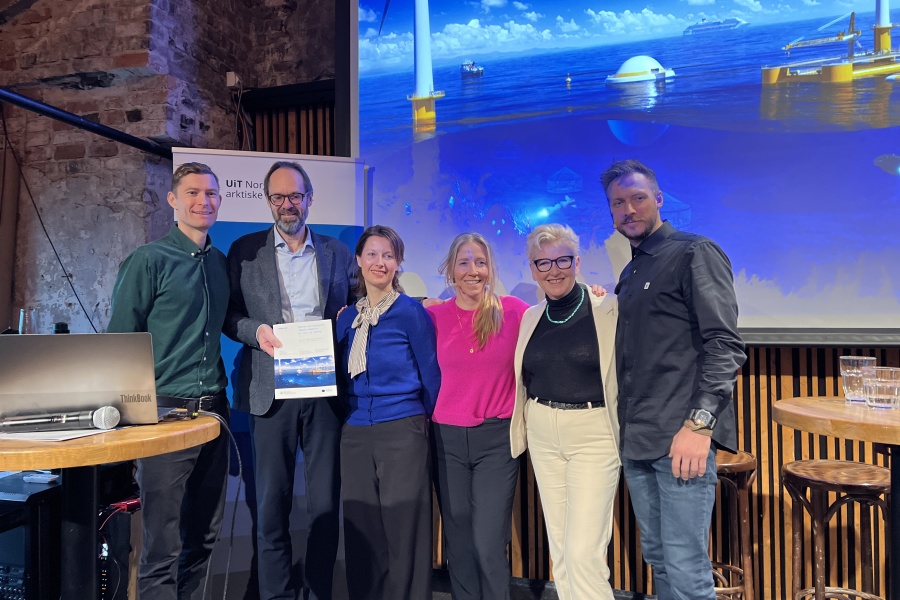 En gruppe på seks personer står samlet på en scene foran et stort lerret som viser en havvind-installasjon. De smiler mot kamera, og en person holder et dokument.