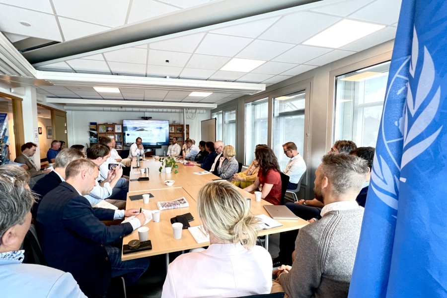 Møte i konferanserom med mange deltakere samlet rundt et langt bord, presentasjon vises på skjerm i enden av rommet, med en FN-flagg synlig i forgrunnen.