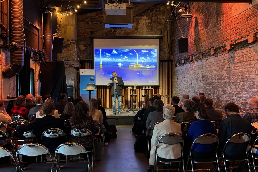 En person står på en scene og holder en presentasjon foran et publikum i en sal med murvegger. Et stort lerret viser et bilde av en marin næringspark.