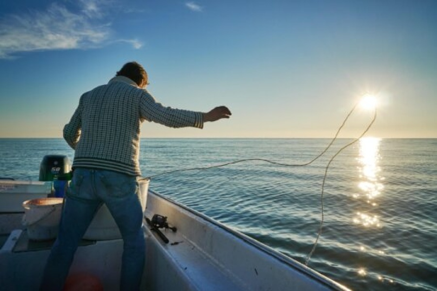 En person står i en liten båt og kaster en line ut i havet mens solen står lavt over horisonten. Sollyset glitrer i det rolige vannet og skaper en varm stemning i kveldssolen.