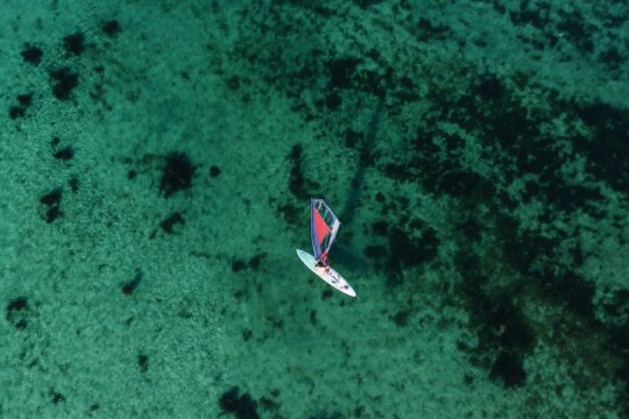 Luftfoto av en person som seiler på et brett med fargerikt seil over grunt, turkist vann. Havbunnen med mørke flekker av tang og stein skimtes tydelig gjennom det klare vannet.