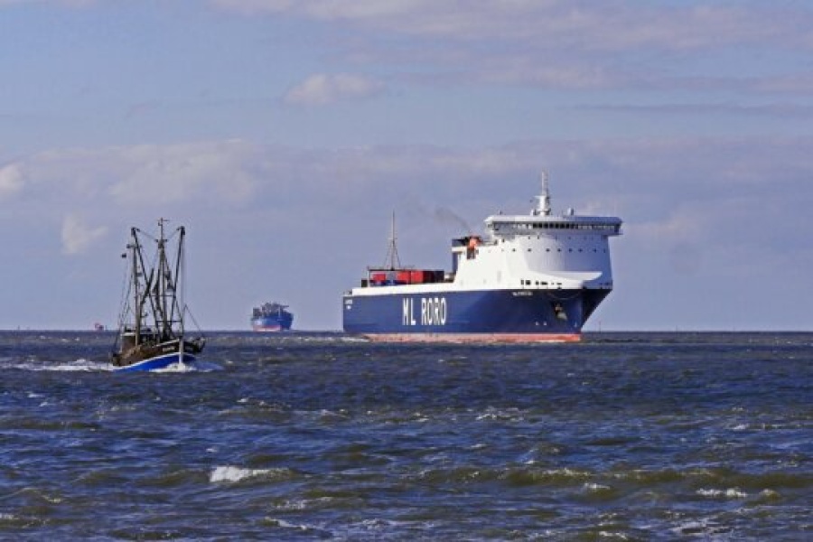 Et stort lasteskip med teksten "ML RORO" på skroget seiler på havet, mens en mindre fiskebåt passerer i forgrunnen. I horisonten skimtes flere fartøy under en delvis skyet himmel.
