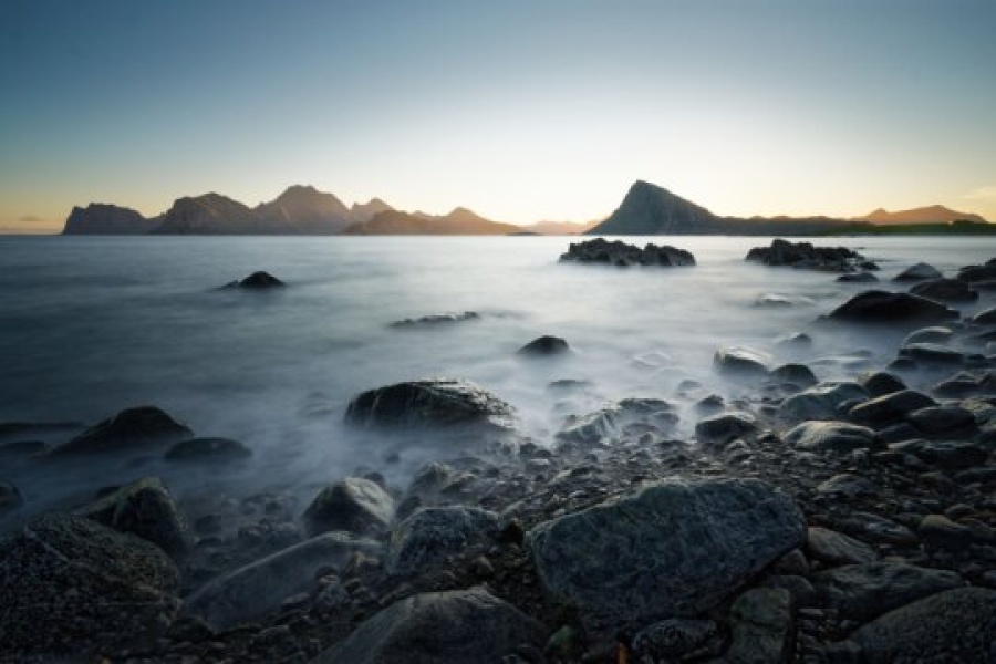 Kystlandskap i solnedgang med steiner i forgrunnen og fjell i horisonten. Lang eksponering gir havet et sløret, tåkete uttrykk som kontrasterer mot de mørke fjellformasjonene i bakgrunnen.