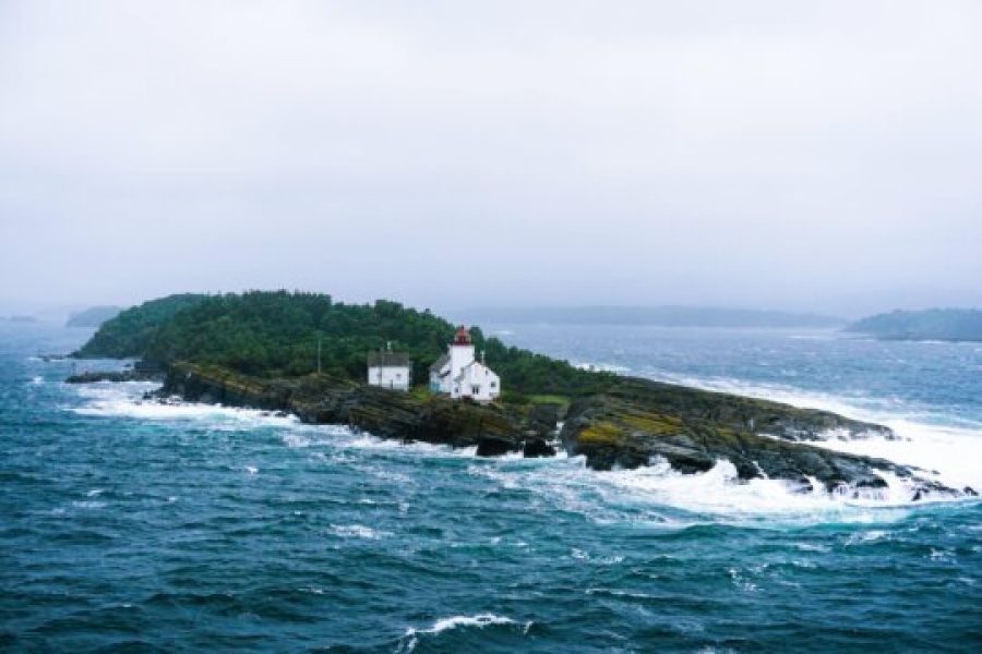 En liten øy med et hvitt fyrtårn og bolighus omgitt av grønn vegetasjon, ligger ute i et opprørt hav. Bølgene slår mot klippene, og tåke og gråvær gir landskapet et dramatisk kystpreg.