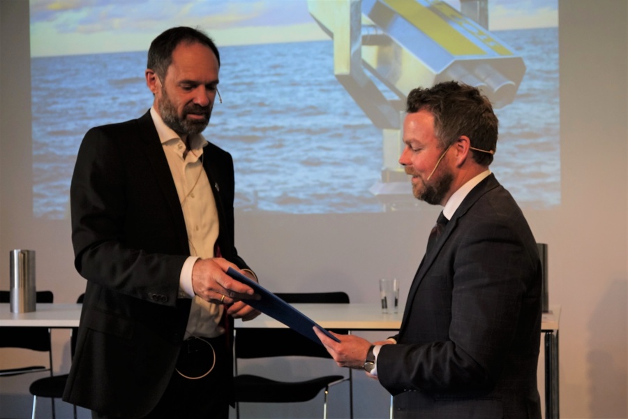 To menn i dress står på en scene under en presentasjon, der den ene overrekker et dokument til den andre. I bakgrunnen vises et bilde av havet og en kikkert på storskjerm.