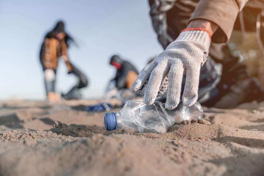 Hånd med hanske plukker opp en plastflaske fra sanden under strandrydding, med flere personer som rydder i bakgrunnen.