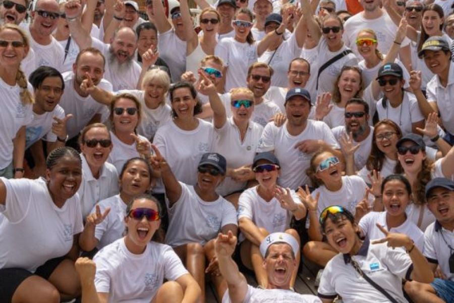 Stort gruppebilde av mennesker i hvite t-skjorter på dekket av et seilskip, smilende og hevende armene i en begeistret pose.