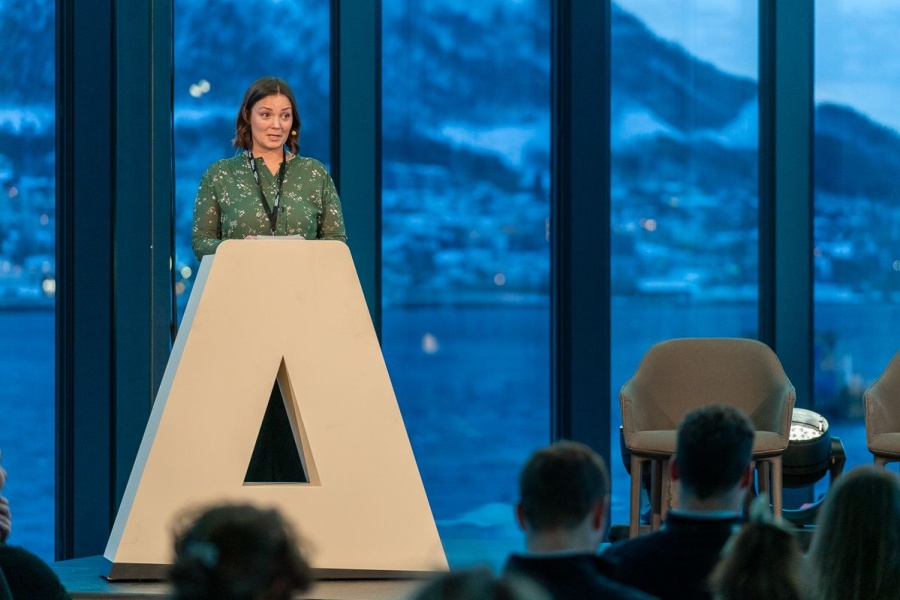 En person står på en scene bak en stor talerstol formet som en “A”, og holder et innlegg foran et publikum, med vinterlandskap og fjell synlig gjennom store vinduer i bakgrunnen.