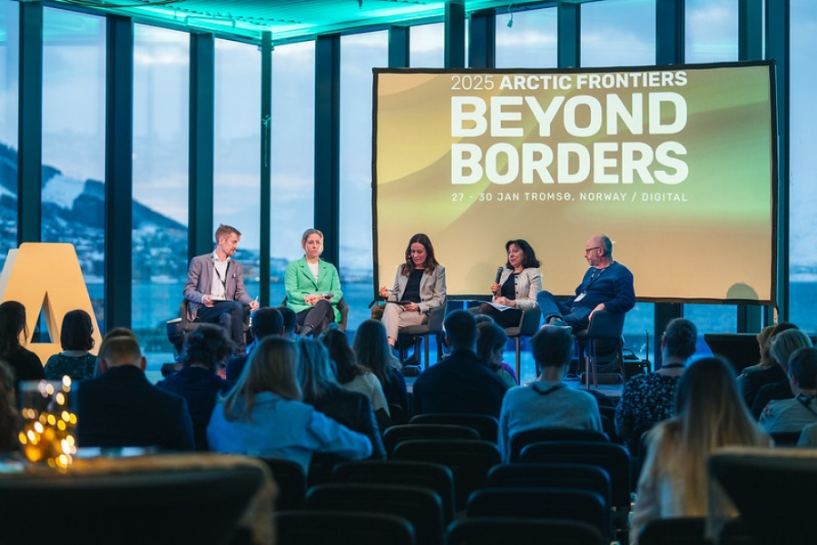 Et panel med fem personer sitter på en scene foran et publikum under Arctic Frontiers 2025, med et stort bakskjermbanner som viser teksten “Beyond Borders” og utsikt over fjell og hav gjennom de store vinduene i bakgrunnen.