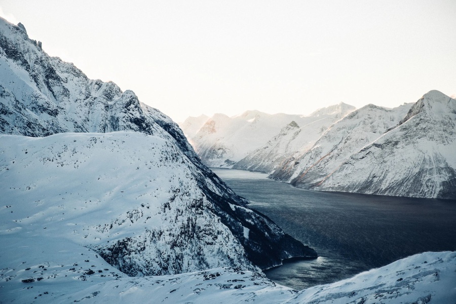 Panoramisk vinterlandskap med en smal fjord som snor seg mellom høye, snødekte fjell under en blek himmel. Solen kaster et mykt lys på fjelltoppene, og mørkeblått vann kontrasterer mot de hvite fjellsidene.