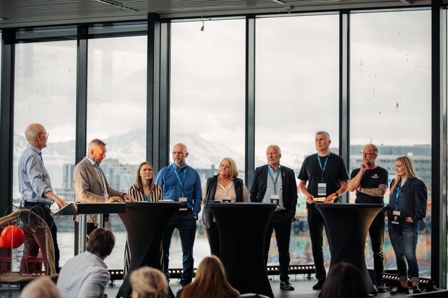 Et panel med ni personer fra ulike deler av sjømatnæringen og forvaltningen står samlet foran store vinduer med utsikt mot fjell og by. De deltar i en panelsamtale foran publikum under en konferanse.