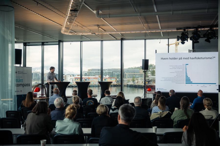 En foredragsholder presenterer for et publikum under et Blått Kompass-arrangement, med en stor skjerm som viser statistikk om havfisketurisme og utsikt mot havn og snødekte fjell gjennom vinduene bak.
