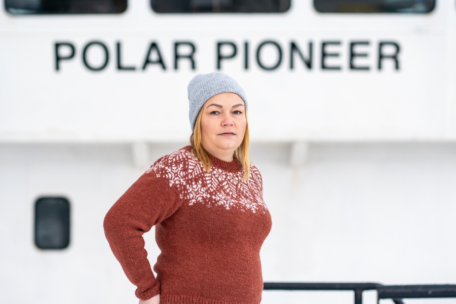 En kvinne med grå strikkelue og brun ullgenser med hvitt mønster står ute foran et skip med navnet «POLAR PIONEER» i bakgrunnen.
