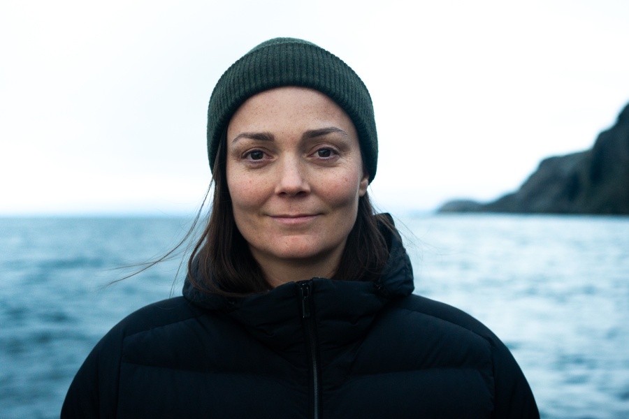 Person iført mørk jakke og grønn lue står utendørs foran et hav og fjell i bakgrunnen på en overskyet dag.