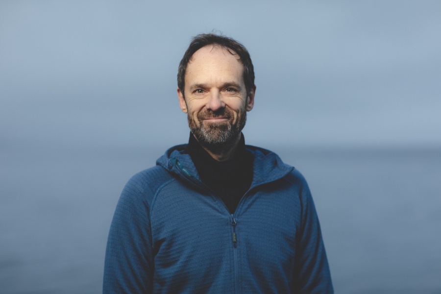 En mann står utendørs foran havet og smiler mot kameraet. Han har på seg en blå hettegenser og har kort hår og skjegg.