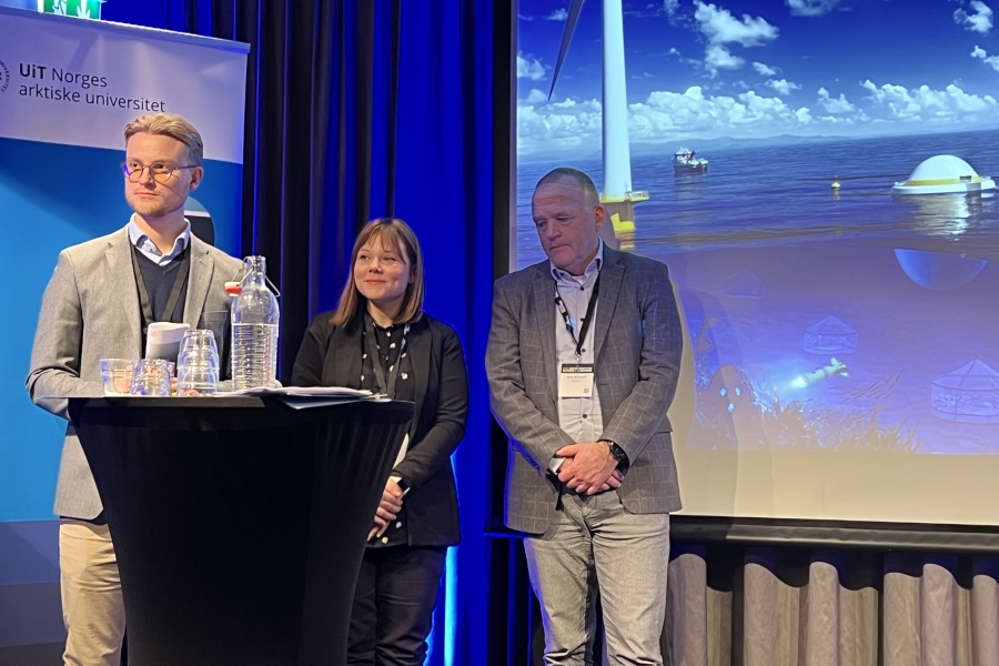 Tre personer står på en scene under et arrangement ved UiT Norges arktiske universitet; en presentasjons­skjerm bak dem viser et havmiljø med offshore vindmølle og havinstallasjoner.