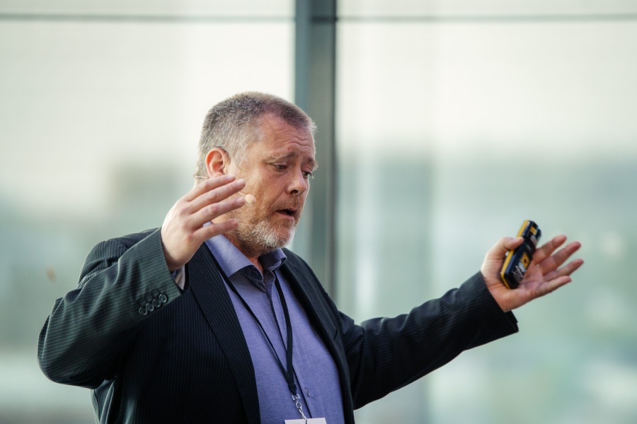 Mann med skjegg og mikrofon gestikulerer engasjert mens han holder en presentasjonsklikker.