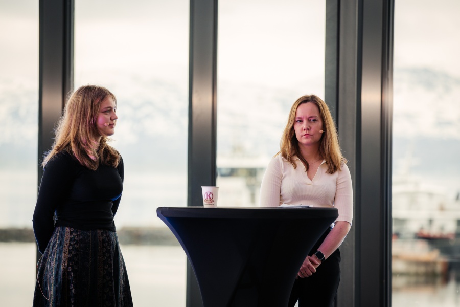 To unge kvinner står på en scene bak et høyt rundt bord under en paneldiskusjon. Begge har mikrofoner og ser konsentrerte ut. I bakgrunnen sees store vinduer med utsikt over snødekte fjell og fjord.