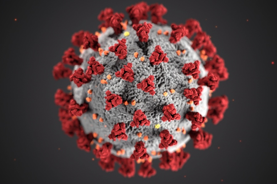 3D illustration of the SARS-CoV-2 virus, showing a spherical gray particle covered with red spike proteins and small orange surface proteins on a dark background.