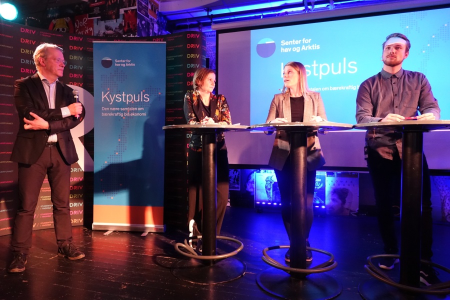 Fire personer deltar i en paneldebatt på en scene under arrangementet «Kystpuls», arrangert av Senter for hav og Arktis. En mann står med mikrofon til venstre, mens tre personer står bak høye bord foran en skjerm med arrangementets logo.