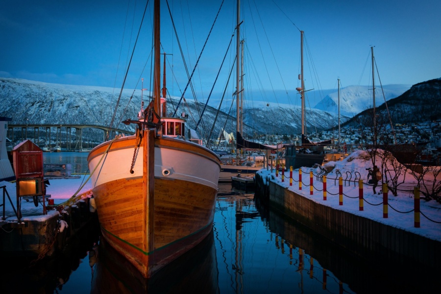 Trebåt fortøyd ved kai i vinterlys i Tromsø havn, med Tromsøbrua og snødekte fjell i bakgrunnen.