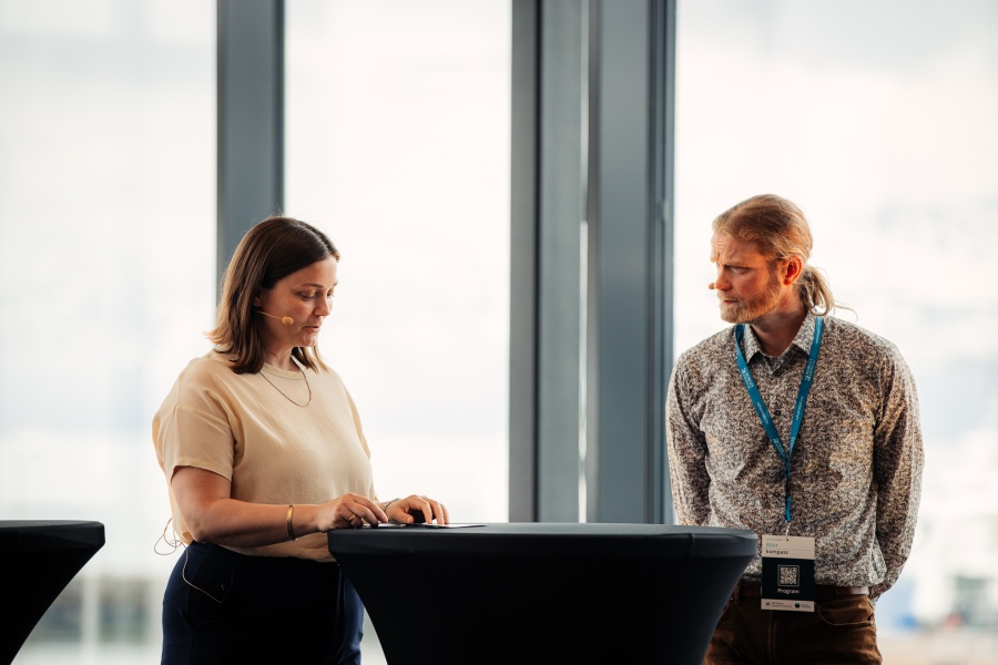 To personer deltar i en panelsamtale foran store vinduer med utsikt over et snødekt fjellandskap. Kvinnen til venstre står bak et rundt bord og ser ned på notatene sine, mens mannen til høyre ser på henne og lytter oppmerksomt. Begge har mikrofoner festet ved kinnet.