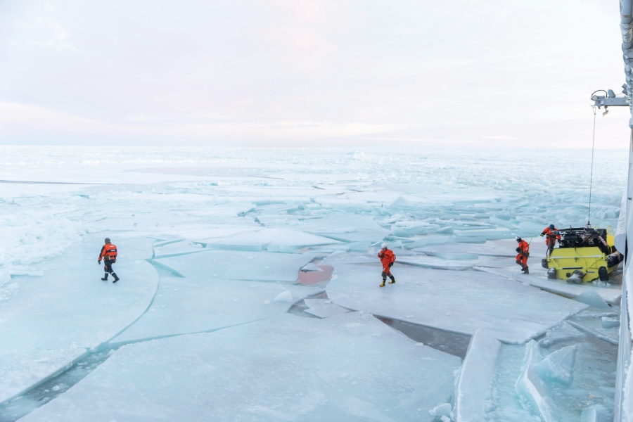 Forskere i oransje overlevelsesdrakter arbeider på isflak i et arktisk landskap, med et gult kjøretøy og et skip ved kanten av bildet.