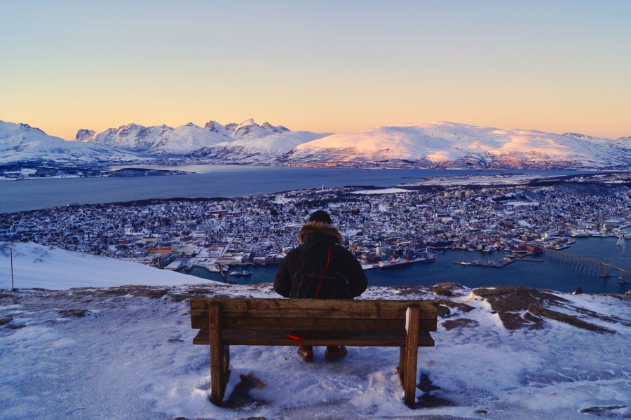 En person sitter på en benk og ser ut over Tromsø by, omgitt av snødekte fjell og hav i solnedgang.
