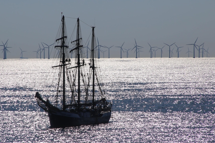 Et seilskip ligger på glitrende hav med en rekke vindmøller til havs i bakgrunnen, som illustrerer kontrasten og sameksistensen mellom tradisjonell sjøfart og moderne fornybar energi.