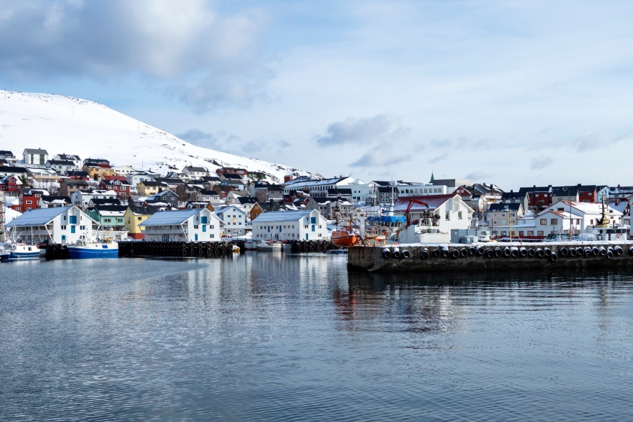 Kystby med fargerike hus og fiskebåter ved kai, omgitt av snødekte fjell og rolig hav i nordnorsk vinterlandskap.